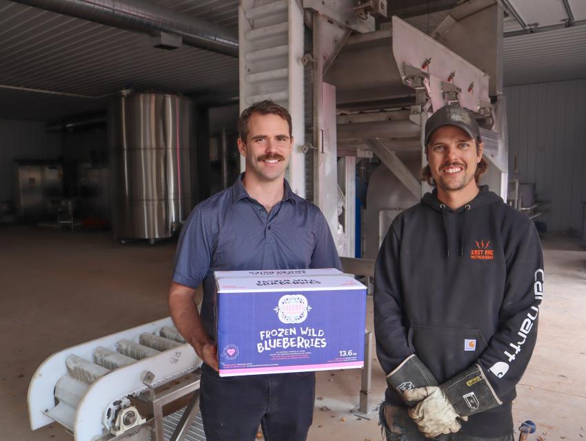 Two men stand in a processing plant, one holding a box of frozen wild blueberries.